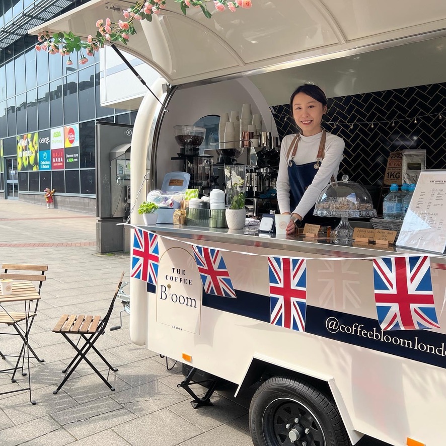 Coffee Bloom Woman stood inside a coffee trailer with cake stand and England bunting